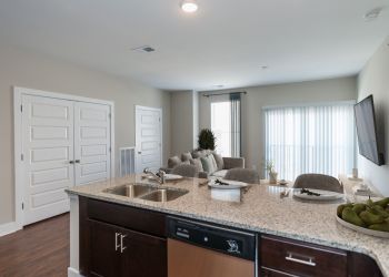 Modern open-plan living room and kitchen with granite countertop, sofa, and large windows with vertical blinds.
