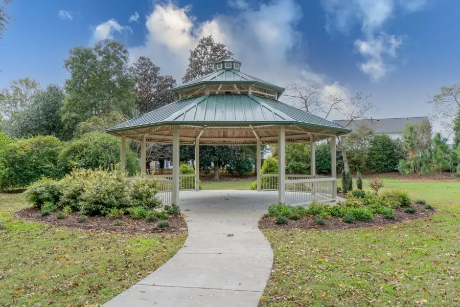 The Amber at Greenbrier A green-roofed gazebo sits in a park, surrounded by bushes and trees under a partly cloudy sky.