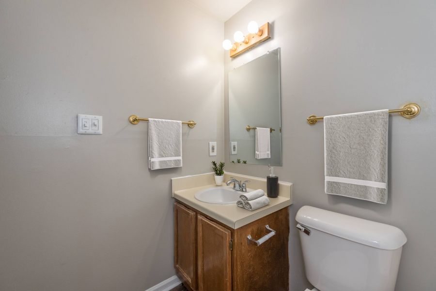 Modern bathroom with a sink, mirror, light fixture, towels, and a toilet against light gray walls.