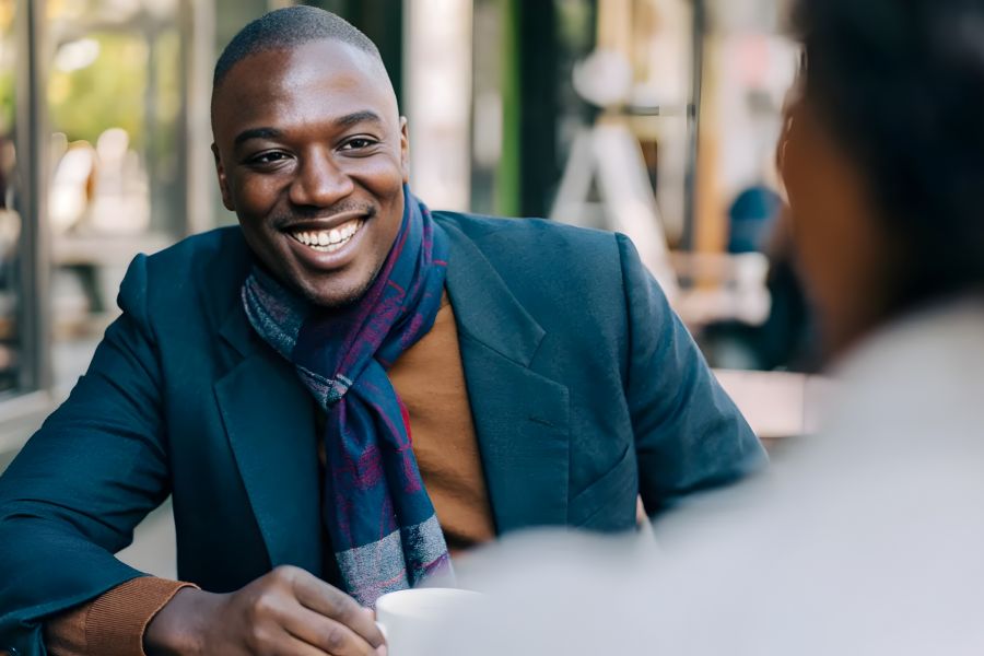 The Amber at Greenbrier Smiling man in a blazer and scarf sits at a table, talking with someone in an outdoor cafe.