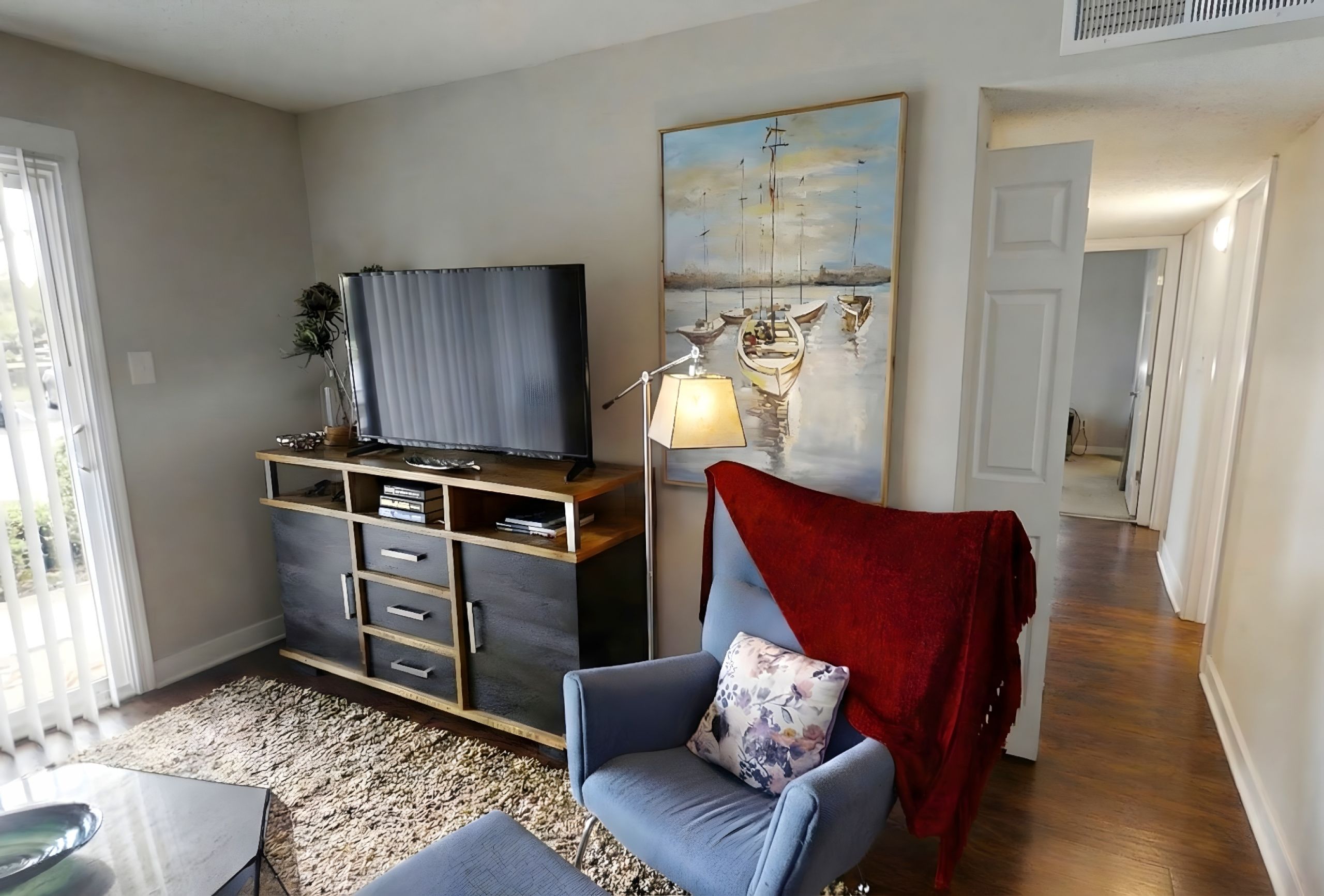 The Amber at Greenbrier A cozy living room with a TV, armchair, red throw blanket, and a painting of boats on the wall.