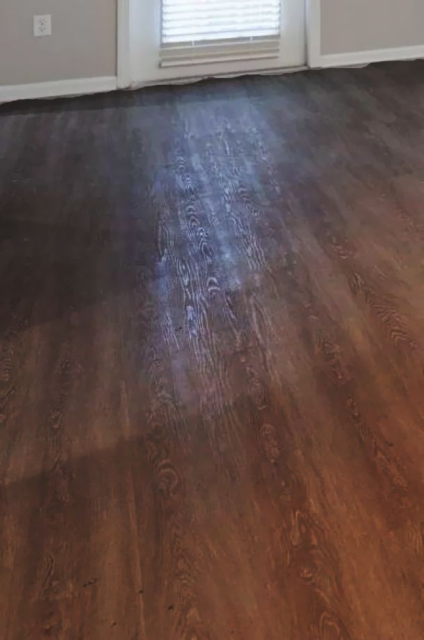 Brown wood-look flooring in a room with beige walls, a white baseboard, and a window with closed blinds.