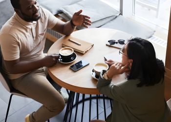 The Amber at Greenbrier Two people sit at a round café table, talking and drinking coffee, with notebooks and a phone on the table.