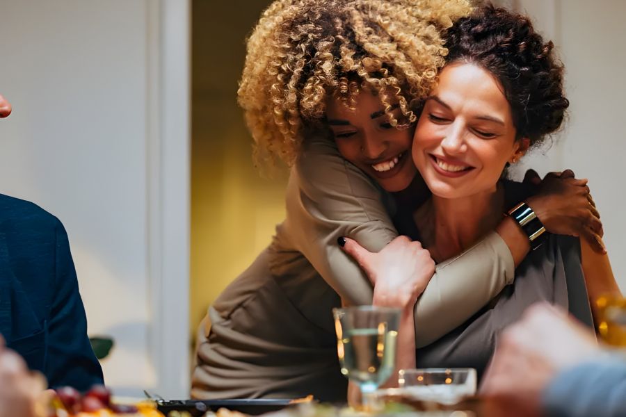 The Amber at Greenbrier Two women smiling and hugging at a dinner table, surrounded by food and drinks, sharing a joyful moment.