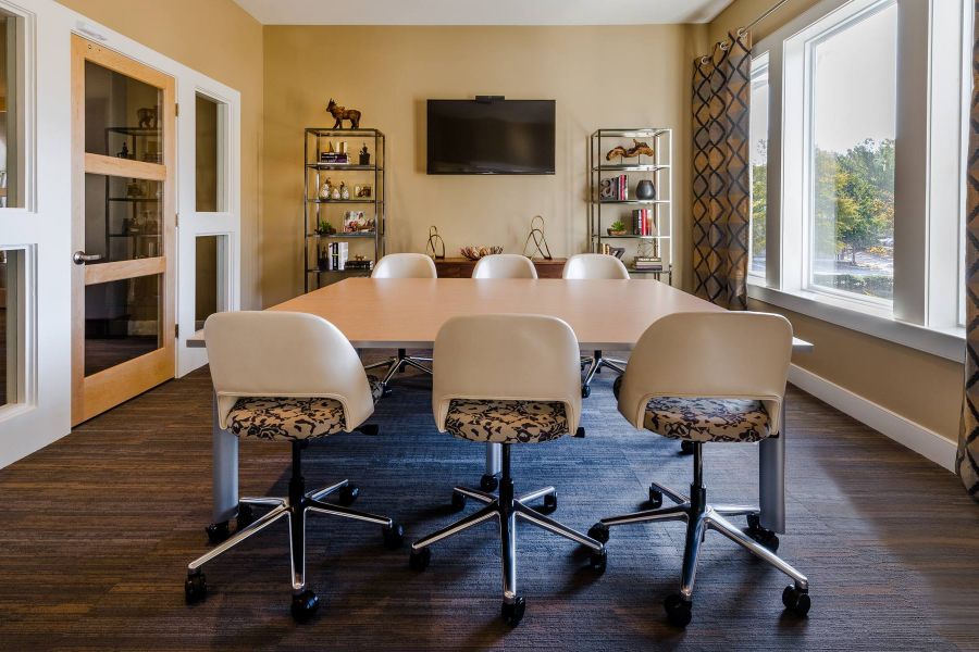 The Amber at Greenbrier Modern meeting room with a large table, six chairs, wall-mounted TV, shelves, and large windows with curtains.